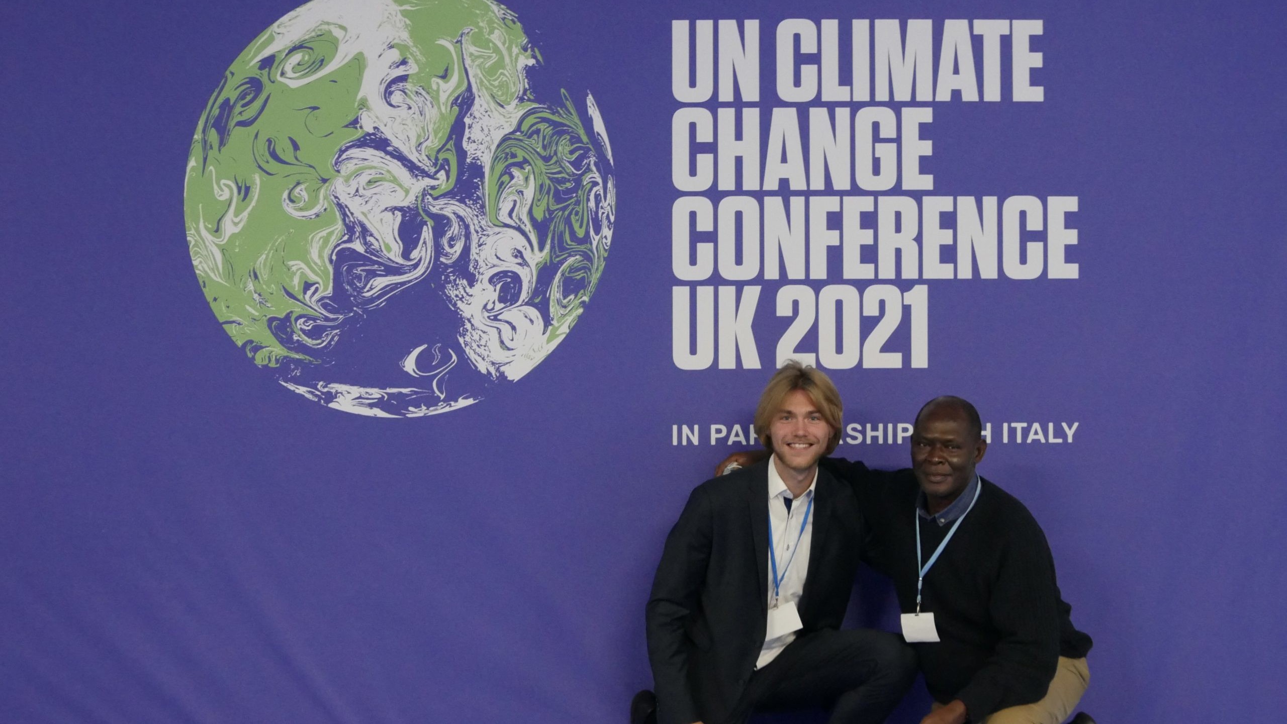 Nico and Fred Onduri kneel in front of COP26 wall