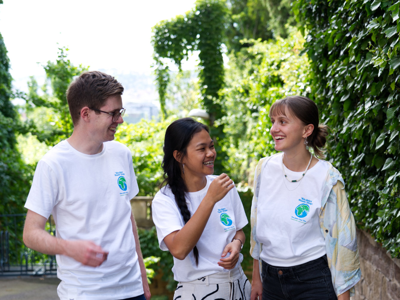 Mathias, Monalisa und Marei tragen das Plant Hope-T-Shirt - auf dem weißen Shirt ist eine blau-grüne Erde und der Schriftzug: This Shirt Plants Hope